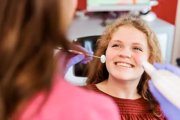 Mädchen sitzt lächelnd beim Zahnarzt, Zahnärztin untersucht die Zähne mit Spiegel.
